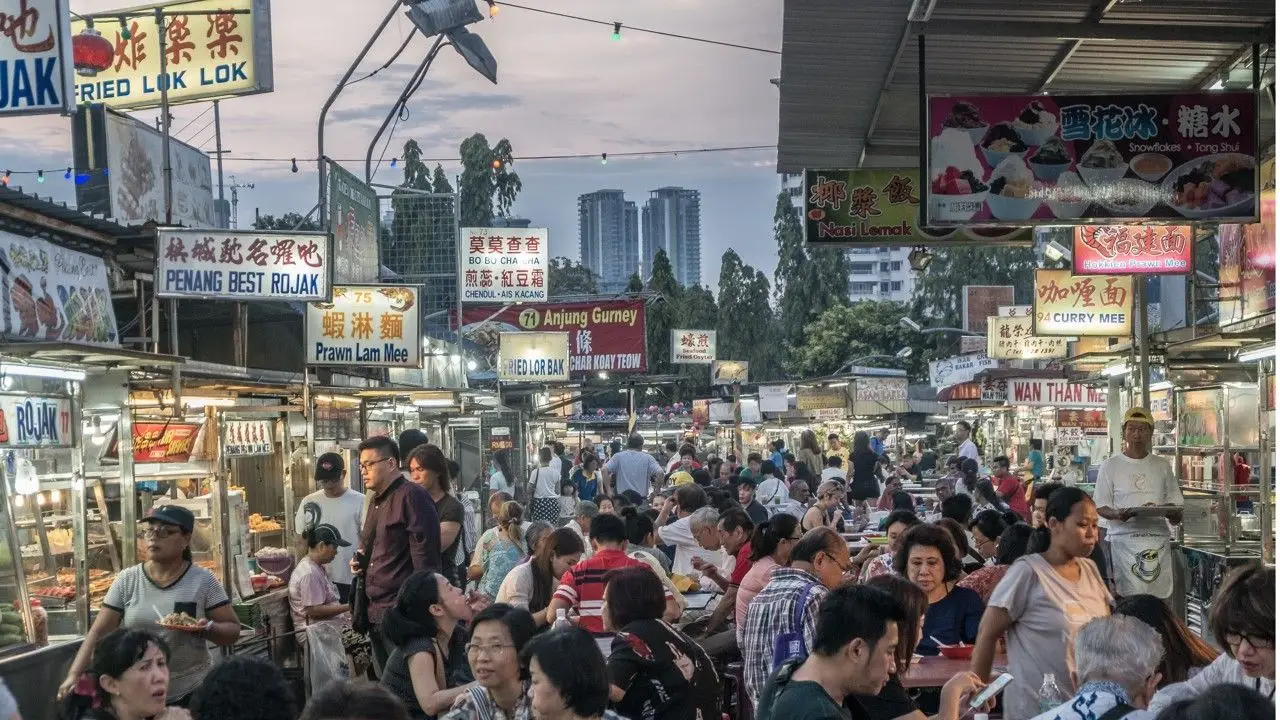 Penang hawker centre at golden hour, packed with locals at long tables under bilingual stall signs.